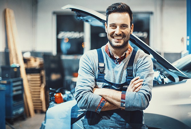 A mechanic smiling with his arms folded standing in front of the side of a vehicle with its hood up.